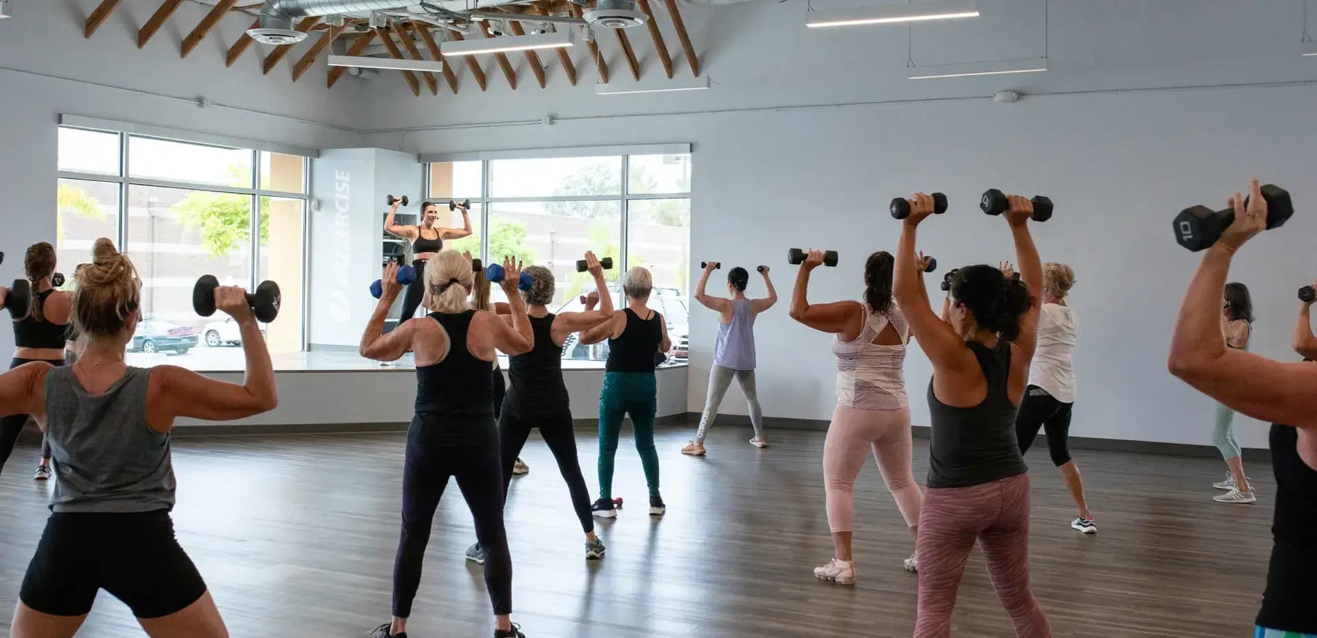A group of people in workout attire lift dumbbells over their heads during a fitness class in a bright, spacious studio with large windows and wooden beam ceiling accents.