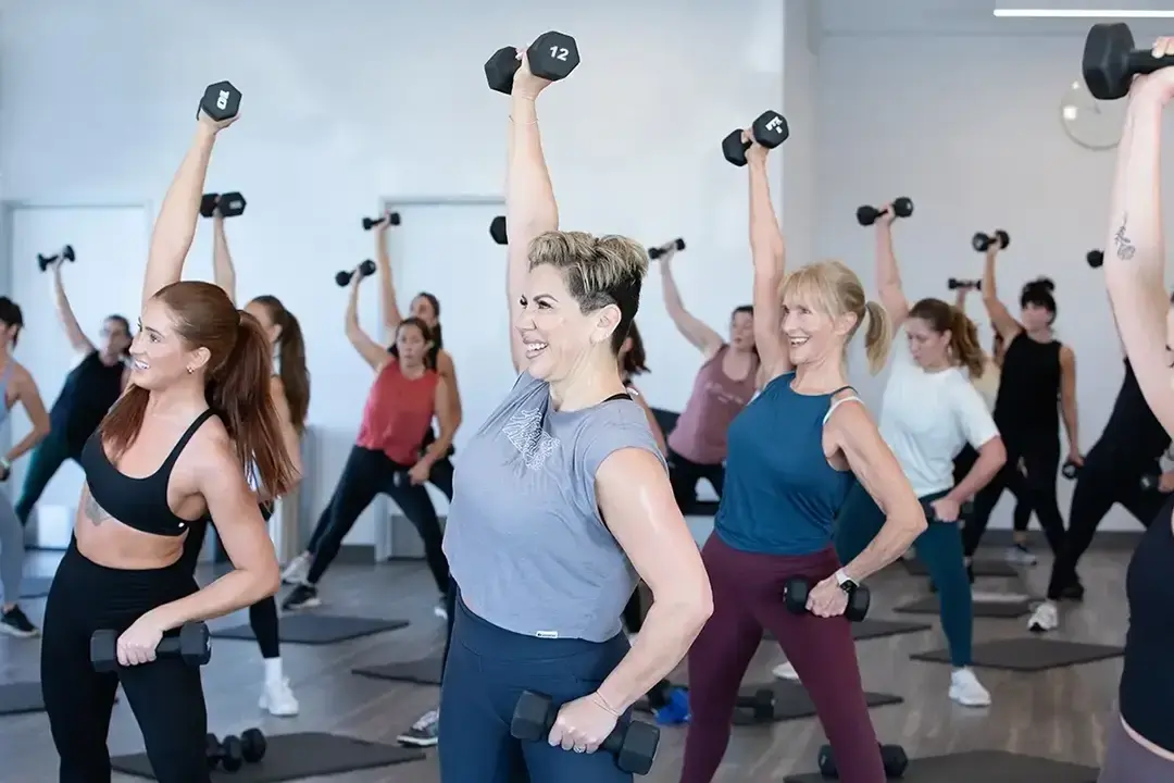 A group of women in athletic wear participate in a fitness class, lifting dumbbells overhead and smiling. They are exercising in a bright, spacious studio with yoga mats on the floor.